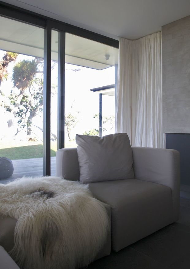 A softly lit minimalist sitting area with a large upholstered armchair, oversized cushion, and a shaggy white throw draped over a low ottoman or seat. Full-height dark-framed glazing opens to a covered outdoor terrace and landscaped garden, while sheer white curtains and a textured pale wall create a calm, neutral interior.