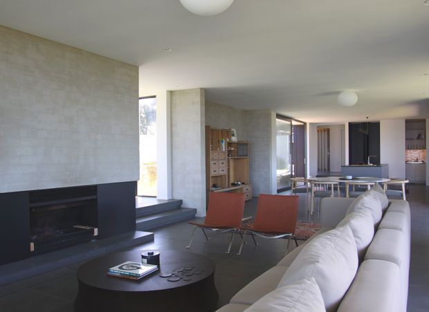 Open-plan contemporary living space with a large black fireplace and hearth at left, a white sectional sofa and round coffee table in the foreground, and two rust-coloured lounge chairs on a rug. The room flows into a dining area and kitchen, with exposed concrete-look walls, dark flooring, extensive glazing, and simple spherical ceiling lights.