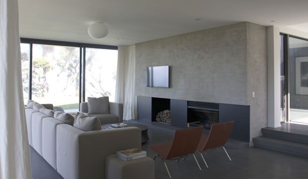 Contemporary living room with a large white sectional sofa, two brown leather lounge chairs, and a minimalist dark built-in fireplace and media wall set against a textured concrete-look feature wall. Floor-to-ceiling glazing and sheer curtains bring in abundant natural light, with steps leading toward an adjacent outdoor area.
