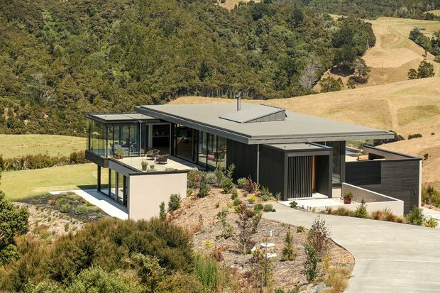 A contemporary single-storey home set into a sloping rural landscape, with a broad low-pitched flat roof, dark cladding, extensive floor-to-ceiling glazing, and projecting glazed living areas. A landscaped driveway and native-style garden surround the house, with rolling pasture and dense bush beyond.