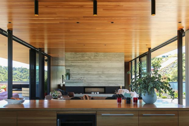 A contemporary open-plan living room viewed across a timber kitchen island or cabinetry. The room features a full-width warm timber ceiling, expansive black-framed glazing, a large pale stone or concrete fireplace wall with a linear built-in fire, dark structural posts, and views toward a green suburban landscape. A dining or living area is visible beyond the island, with abundant natural light and indoor plants.