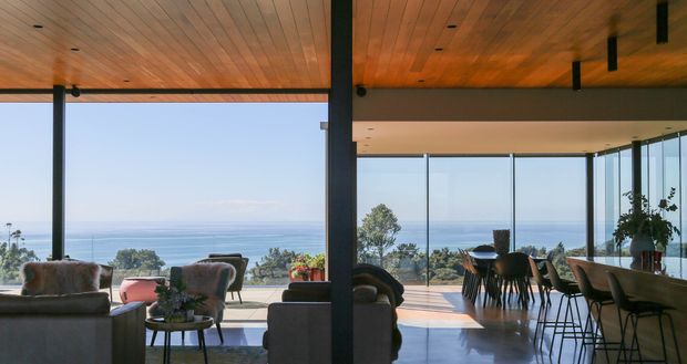 An open-plan coastal living and dining space with expansive floor-to-ceiling glazing overlooking the ocean. The room features a warm timber-lined ceiling, dark structural posts and window frames, polished light flooring, a lounge seating area, a dining table, and a kitchen or bar counter with stools. The bright, airy interior connects directly to an ocean-facing terrace.