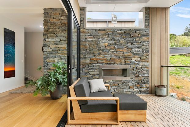 Contemporary indoor-outdoor living area with a floor-to-ceiling glazed opening connecting a warm timber-floored interior to a timber deck. A stacked natural stone feature wall incorporates a recessed fireplace, while a timber-framed lounge chair and ottoman sit beside potted greenery. Vertical timber wall cladding and expansive glazing create a bright, refined connection to the landscaped exterior.