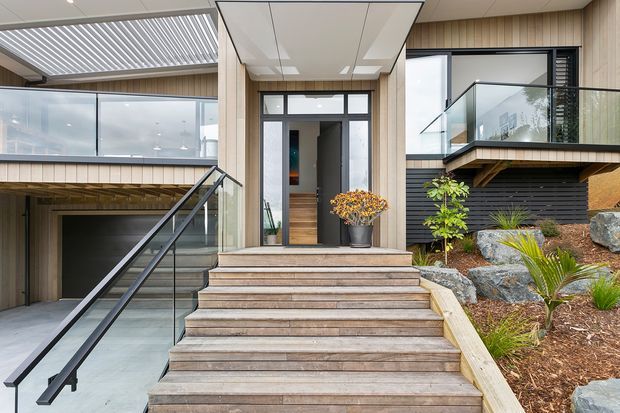 Contemporary residential entrance viewed from the front, with a broad timber staircase leading to a glazed entry door beneath a covered canopy. The facade combines vertical timber-look cladding, dark horizontal cladding, expansive glazing, black metal-framed balustrades, and landscaped rock garden beds. A lower-level garage or undercroft is visible to the left.