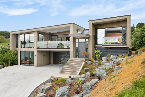 Exterior view of a contemporary multi-level hillside house with a rectilinear stepped form, light vertical timber-look cladding, dark window surrounds, extensive glazing, a glazed balcony, and an integrated lower-level garage. Broad concrete driveway and entry stairs lead through a landscaped rocky slope.