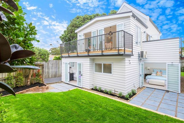 A bright two-storey residential dwelling with white horizontal weatherboard cladding, a pitched gabled roof, and a projecting upper-level balcony with dark metal balustrades. The lower level opens onto a landscaped lawn and paved paths, with a garage or covered parking area at right and a visible bedroom or living space inside. The scene has a clean suburban coastal feel with strong blue-sky daylight.
