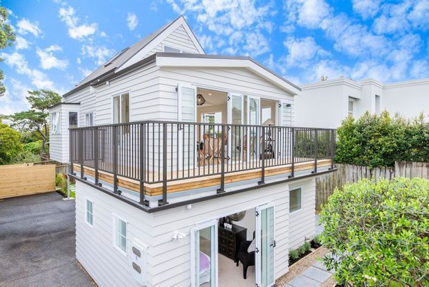 Exterior view of a white weatherboard, multi-level home with a prominent gabled roof, upper-level timber deck and dark vertical metal balustrade. Large glazed doors open from the upper floor onto the deck, while the lower level has additional access doors and windows. Dense greenery surrounds the property.