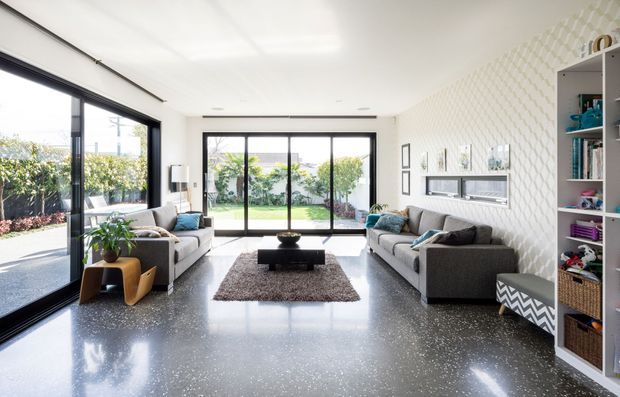Bright contemporary living room with full-height black-framed glazing opening to a garden, a polished dark terrazzo floor, grey sofas, a central coffee table and rug, built-in wall shelving, and a textured geometric feature wall with a recessed fireplace or media niche.