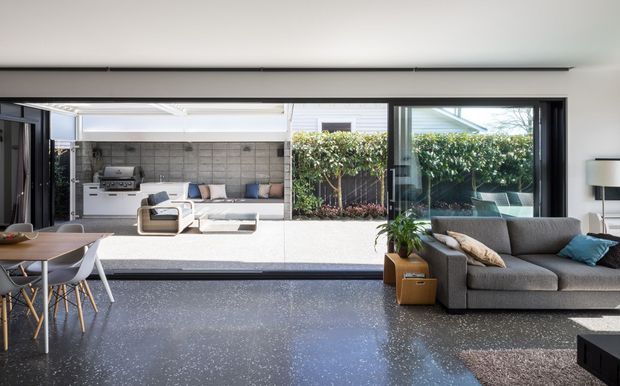 Contemporary open-plan living and dining area with a dark polished terrazzo-style floor, streamlined built-in glazing, and wide sliding doors opening to a landscaped courtyard and covered outdoor entertaining area. A grey sectional sofa, dining table, and neutral furnishings create a relaxed indoor-outdoor connection, with an outdoor grill visible beyond.