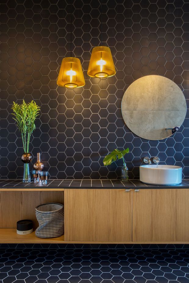 A moody, refined bathroom vanity area featuring dark hexagonal wall and floor tiles, a floating timber vanity with integrated storage, a pale round vessel basin, a large circular mirror, and two warm amber pendant lights. Decorative plants and accessories add organic contrast.