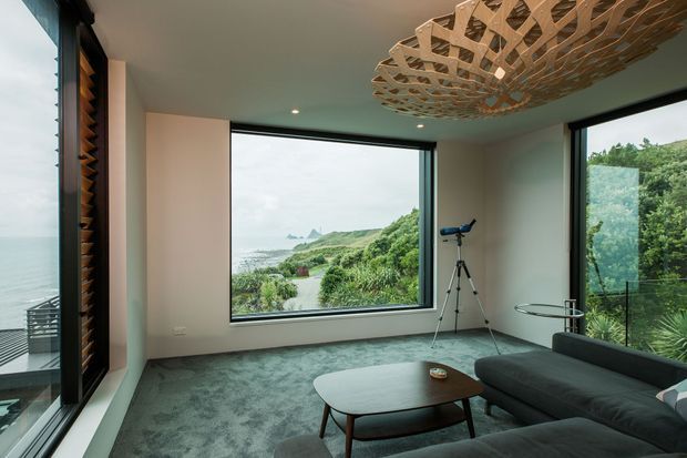 A contemporary living room with expansive corner glazing overlooking a lush coastal landscape and ocean. The room features a dark sectional sofa, low oval coffee table, grey carpet, pale walls, a dark ceiling, and a sculptural timber ceiling pendant. A telescope is positioned beside the large window.