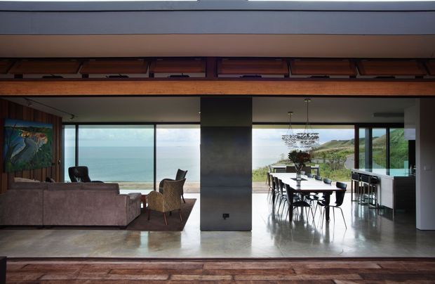 Open-plan coastal living and dining area with expansive floor-to-ceiling glazing overlooking the ocean and a green hillside. The room features a low upholstered sofa, woven lounge chair, long dining table with dark chairs, polished concrete flooring, dark structural cabinetry or wall panels, and a dramatic timber ceiling with exposed beams and built-in clerestory-like storage panels. The palette is warm and muted, with strong indoor-outdoor visual connection and abundant natural light.