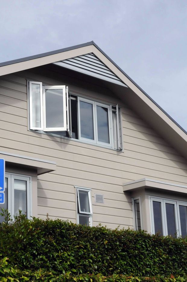 Exterior view of a contemporary residential building with a steep gabled roof, horizontal beige cladding, dark roof edging, and multiple white-framed windows. The upper window includes outward-opening casements, while a neatly trimmed hedge partially obscures the lower level.