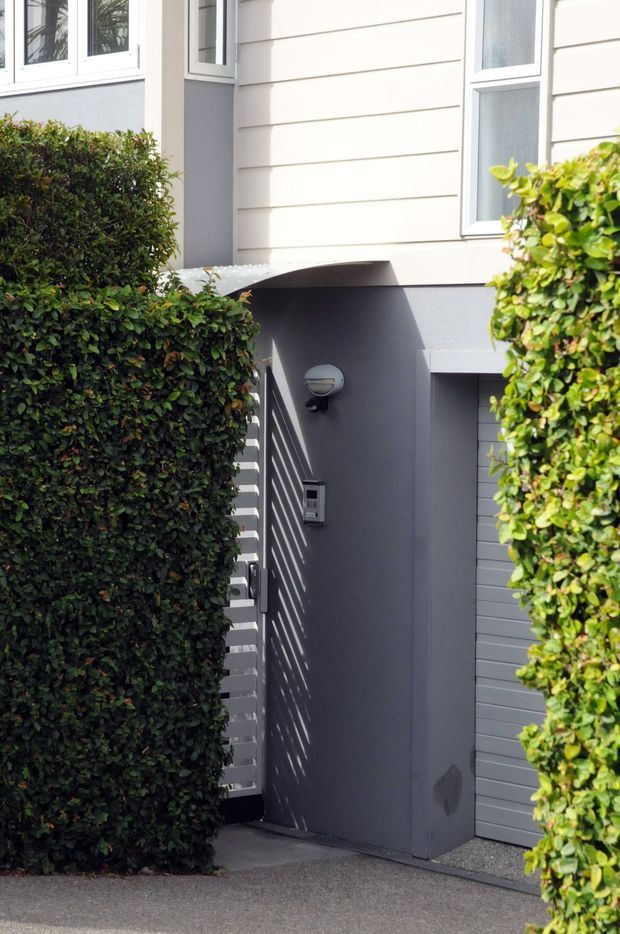 A close exterior view of a contemporary residential entrance partially screened by neatly trimmed hedges. The facade combines light horizontal weatherboard cladding, grey painted rendered walls, white-framed windows, and a recessed entry with a grey garage-style door and wall-mounted light.