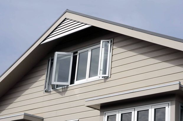Close-up exterior view of a gable-front house with horizontal light-coloured cladding, white-framed windows, and a projecting roof over the entrance. The open casement windows and strong roofline are the dominant features against an overcast sky.