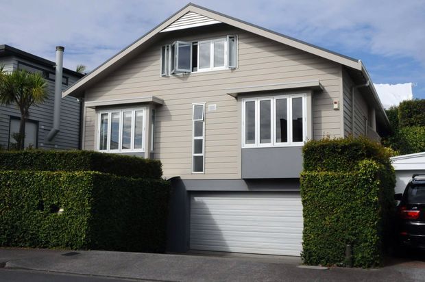 Exterior view of a compact two-storey suburban house with a steep gabled roof, light horizontal weatherboard cladding, white-framed windows, and an integrated lower-level single garage. Carefully trimmed hedges frame the facade and driveway.