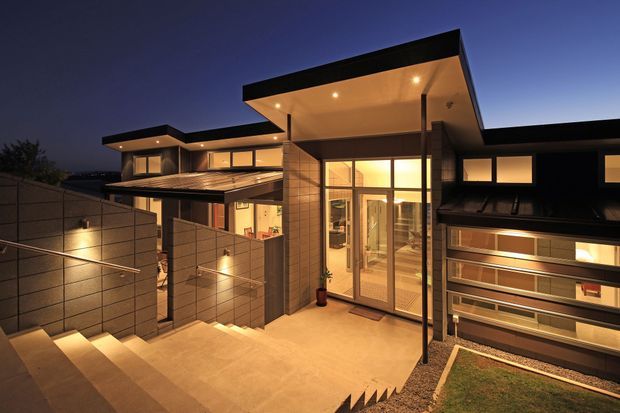 Twilight exterior view of a contemporary multi-level residence with strong horizontal roof planes, warm recessed lighting, extensive glazing, a sheltered entry courtyard, broad steps, and textured dark cladding. The composition highlights the facade, entry approach, and layered roof forms.