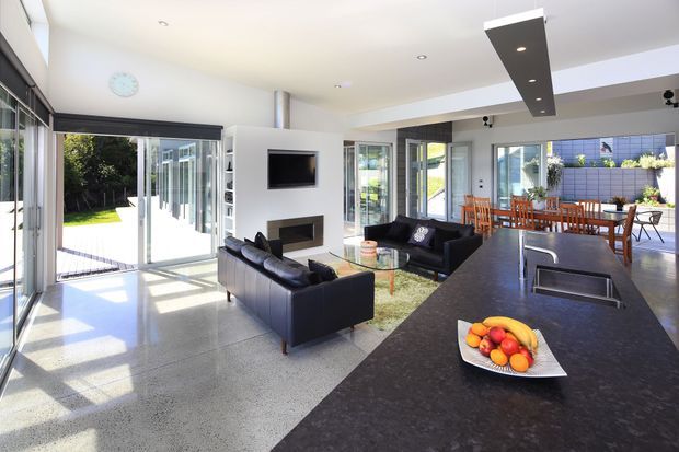 Bright open-plan contemporary living, dining and kitchen area with extensive floor-to-ceiling glazing, polished concrete flooring, a dark stone kitchen island, black sofas, a central fireplace and television wall, and a timber dining setting opening to a landscaped outdoor area.