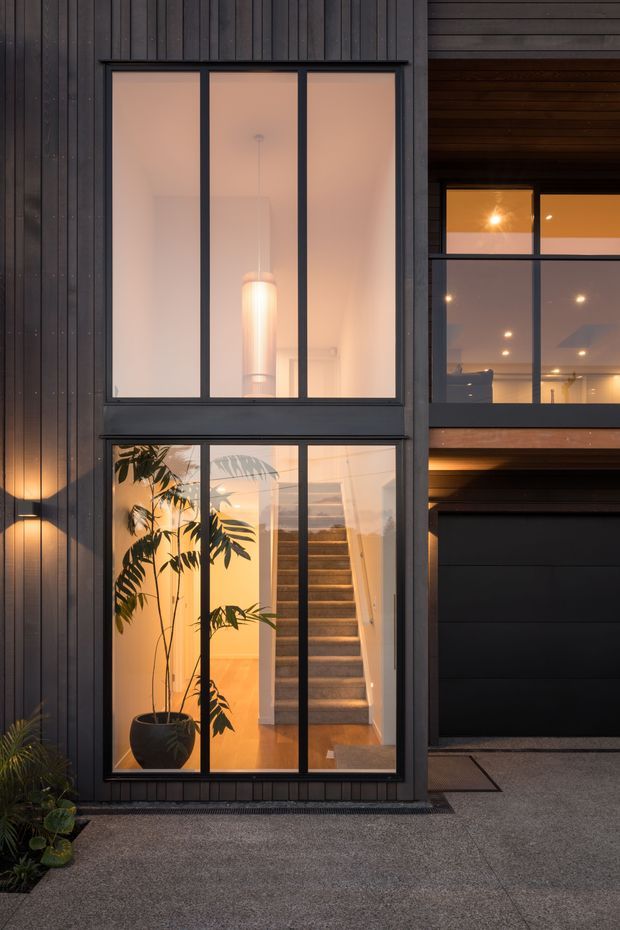 A dusk exterior view of a contemporary multi-level home with dark vertical cladding, black-framed floor-to-ceiling glazing, and a recessed garage. Warm interior lighting reveals a staircase, pendant light, and indoor plants through the windows. The facade has a strong vertical composition and an adjacent covered upper-level opening.