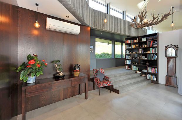 Bright contemporary interior entry or living space with a dramatic double-height ceiling and clerestory glazing. A dark timber feature wall incorporates a floating console and pendant lighting, while broad steps lead to a raised library area with built-in dark shelving. The space includes a patterned armchair, indoor plants, a tall traditional clock, and a sculptural antler chandelier, creating a mix of contemporary architecture and eclectic decorative elements.