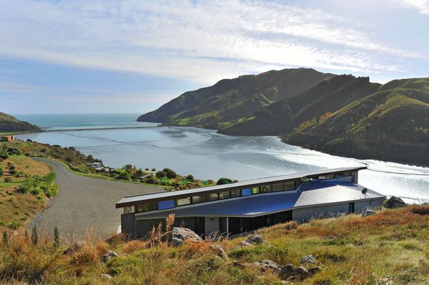 A modern low-profile building with a long dark facade and angular blue-grey roof, set on a grassy coastal hillside overlooking a sheltered bay and rugged green headland. The image is a wide exterior architectural view in bright natural daylight.