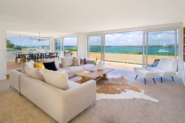 Bright open-plan living and dining area with expansive floor-to-ceiling glazing overlooking a blue waterfront. The room features cream upholstered sofas, sculptural lounge chairs, a timber coffee table, a large hide rug, and a dining table opening onto a timber deck. The interior is airy and contemporary with a strong connection to the outdoor view.