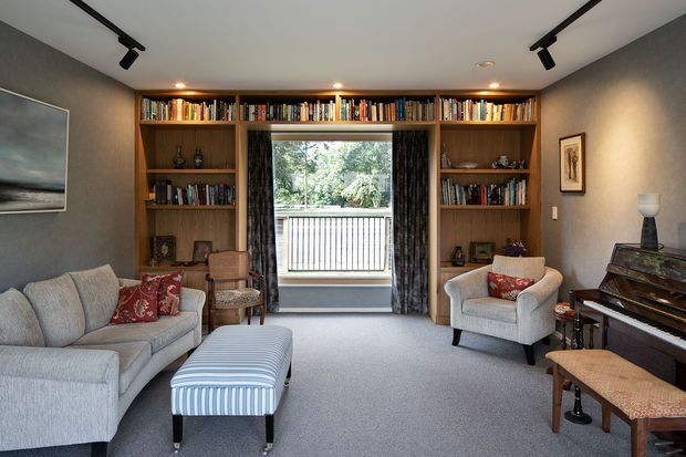 A refined, cozy living room and reading space with full-height dark timber built-in bookshelves framing a central window and balcony view. The room has grey carpet, charcoal textured walls, a pale sectional sofa, upholstered armchair, striped ottoman, upright piano, patterned cushions, and black ceiling track lighting.