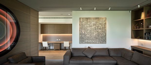 Contemporary living room with a large dark sectional sofa, sculptural wall artwork, built-in shelving and cabinetry, and a recessed study or dining workspace visible beyond. The room features warm timber wall panelling, integrated lighting, neutral finishes, and a refined open-plan layout.
