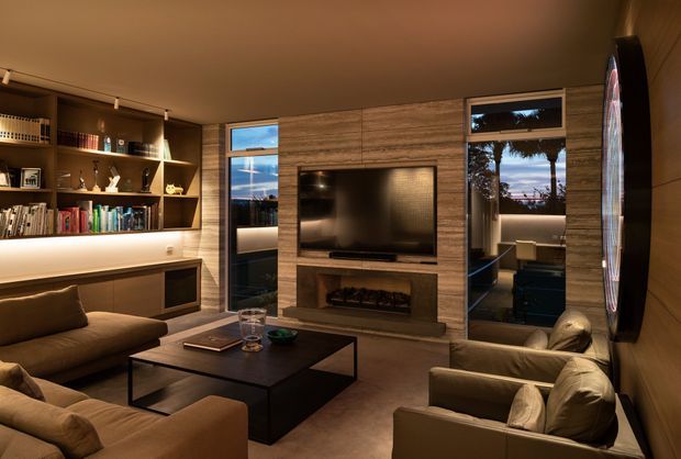 Warm, sophisticated living room with a built-in timber entertainment wall, recessed television, linear fireplace, open shelving, low cabinetry, and large glazing. Neutral upholstered sofas and armchairs surround a dark rectangular coffee table, while reflected outdoor light and sunset views add depth. The room has a refined contemporary feel with integrated joinery and subdued ambient lighting.