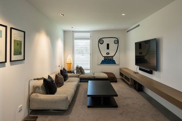 A compact contemporary living room with a large L-shaped upholstered sectional sofa, low black coffee table, and wall-mounted television on a long timber media console. White walls, recessed downlights, carpeted flooring, framed artwork, a roller blind, and playful graphic artwork create a clean, modern interior.