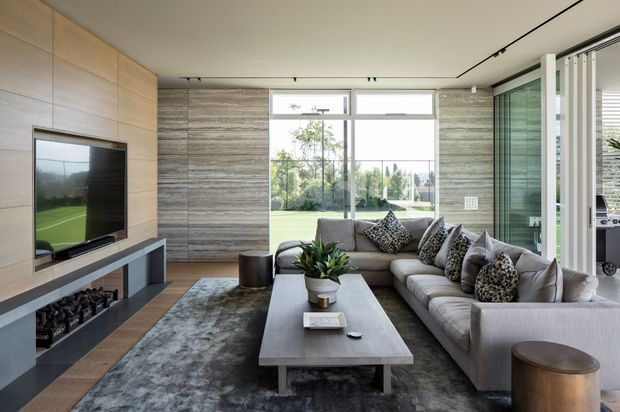 Contemporary living room with a large grey sectional sofa, textured accent cushions, a substantial rectangular coffee table, and a grey area rug. A timber-lined media wall with fireplace sits to the left, while full-height glazing and a sliding glass door bring in abundant natural light. The room combines warm timber, grey stone-like wall cladding, and minimalist recessed lighting.