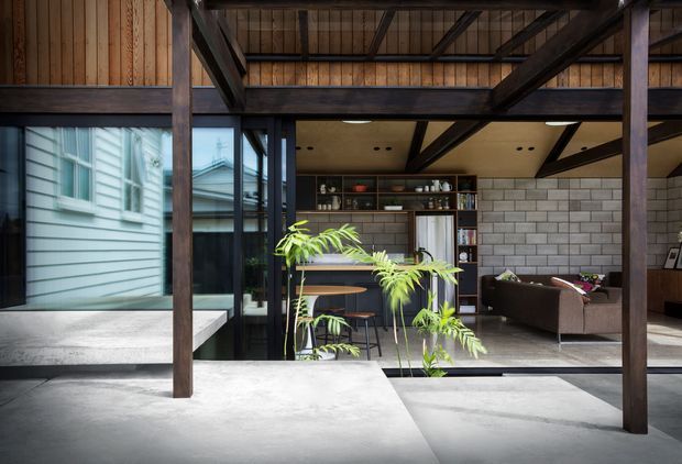 A dramatic open-plan interior framed by dark timber post-and-beam structure, with exposed roof beams, concrete floors and a polished concrete island or platform in the foreground. Glazing opens toward a light weatherboard exterior, while the interior contains a compact kitchen and dining area with timber cabinetry, shelving, stools and a dark sofa. Concrete block walls and warm timber cladding create an industrial yet warm, contemporary character, with a tall indoor plant adding greenery.