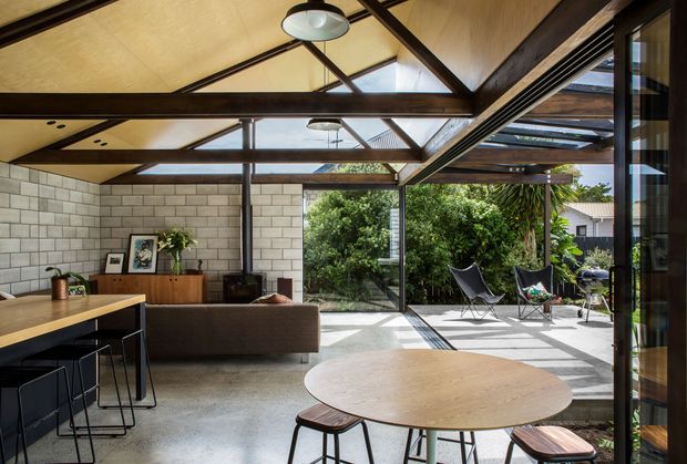 A bright open-plan living and dining area with a compact kitchen island, dining table, sofa, and extensive glazed openings connecting to a paved outdoor terrace. The room features exposed dark timber roof beams, a timber-lined pitched ceiling with skylights, concrete flooring, pale concrete block walls, and strong indoor-outdoor views to greenery. The furniture and finishes create a warm, contemporary mid-century-influenced atmosphere.
