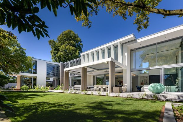A large contemporary residence viewed from a landscaped lawn, featuring crisp white geometric volumes, extensive floor-to-ceiling glazing, a covered outdoor entertaining area, horizontal louvres, and a flat roofline. Mature trees and manicured garden planting frame the house.