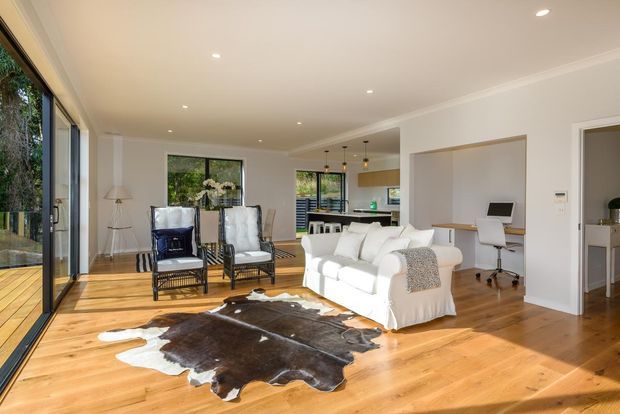 Bright open-plan living room with warm timber flooring, white upholstered sofas, two black-framed lounge chairs, a cowhide rug, and expansive floor-to-ceiling glazing opening toward an outdoor deck. A compact kitchen and a small home office nook are visible in the background. The room has a clean, contemporary residential fit-out with recessed lighting and abundant natural light.