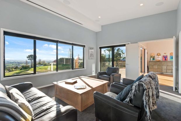 Bright contemporary living room with large picture windows overlooking a landscaped suburban setting. The room features grey walls, dark carpet, black leather sofas and armchairs, a substantial timber coffee table, recessed ceiling lighting, and an open doorway to an adjacent tiled area.