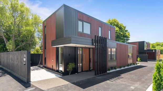 Contemporary multi-level residential building with a strong geometric form, clad in dark vertical metal and warm reddish timber-toned panels. The facade features projecting upper volumes, large windows, a covered entry, and a vertical slatted screen. Paved paths and driveway surround the building, with mature landscaping and a timber boundary fence.