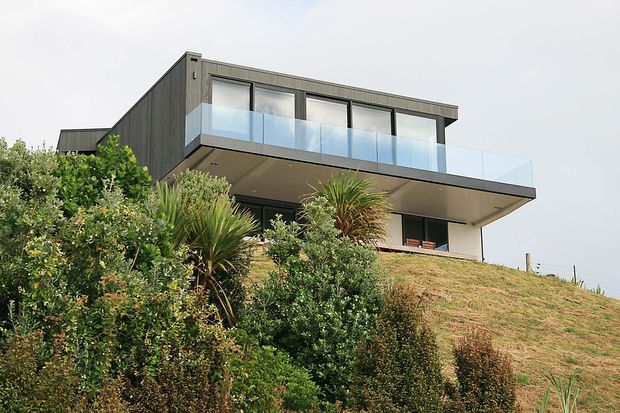 A contemporary two-storey house positioned on a sloping, landscaped site. The building has dark vertical cladding, large upper-level glazing with transparent glass balustrades, broad cantilevered roof and balcony planes, and a light rendered lower facade. Dense garden planting partially screens the lower level.