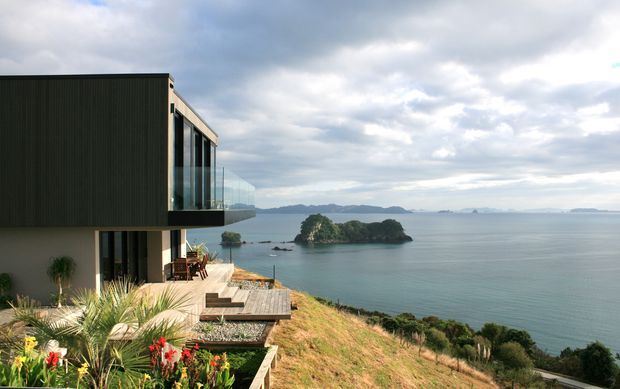Exterior view of a contemporary multi-level coastal home perched on a sloping site above calm blue water. The building has dark vertically ribbed cladding, pale lower walls, extensive floor-to-ceiling glazing, a projecting upper volume, and timber decks. A small island and distant coastline are visible beyond the property, with landscaped planting in the foreground.
