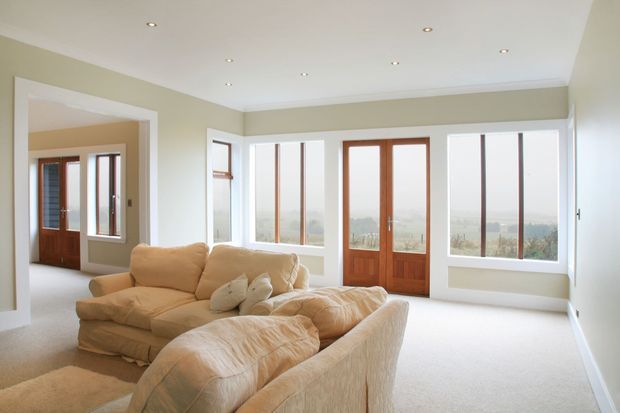 Bright, spacious living room with large floor-to-ceiling windows and timber-framed glazed doors overlooking a distant landscape. The room features pale neutral walls, recessed ceiling lights, light carpet, and a large cream upholstered sectional sofa.