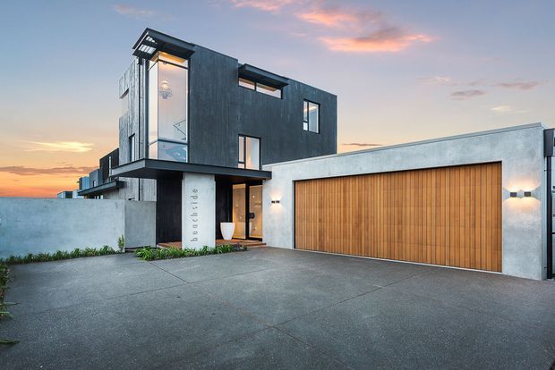 A contemporary multi-level residence viewed from the front at dusk. The facade combines dark vertical cladding, pale rendered walls, extensive glazing, projecting box forms, and a large timber garage door. A broad concrete driveway and entry path lead to the recessed front entrance, with subtle exterior wall lighting.