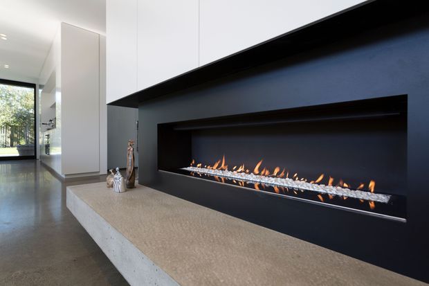 Contemporary minimalist interior centered on a long linear gas fireplace set into a dramatic dark feature wall. A broad pale stone or concrete hearth projects into the room, with crisp white built-in cabinetry above and beside it. Polished grey flooring, clean architectural lines, and glazing visible at the rear create a refined, open living-space atmosphere.