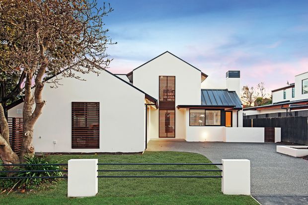 Front exterior view of a contemporary multi-volume residence with crisp white rendered walls, dark metal pitched roofs, timber louvred window screens, a gravel driveway, and a minimalist lawn and entry fence.