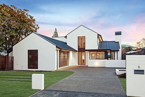 Contemporary multi-wing residential house viewed from the street, with crisp white rendered walls, dark metal pitched roofs, warm timber louvre screens, a garage, paved driveway, lawn, and a minimalist white mailbox in the foreground.