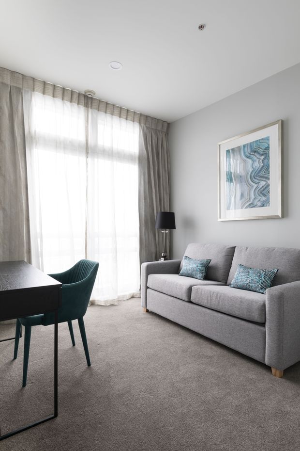 A bright, compact living room with a grey upholstered sofa, teal accent cushions, a dark desk or dining table with a teal chair, floor-to-ceiling sheer curtains and grey drapes, grey carpet, and a framed blue abstract artwork. The room has a simple contemporary fit-out with soft neutral tones and subtle teal accents.