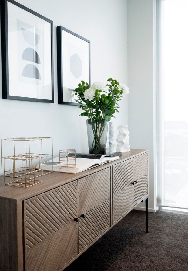 A bright, refined interior vignette featuring a mid-century-inspired timber sideboard with geometric carved door fronts and slim dark legs. The sideboard is styled with a glass vase of greenery and white flowers, decorative objects, books, and brass geometric sculptures. White walls display black-framed abstract artwork, while a full-height glazed opening brings in abundant daylight. Dark carpet provides contrast.