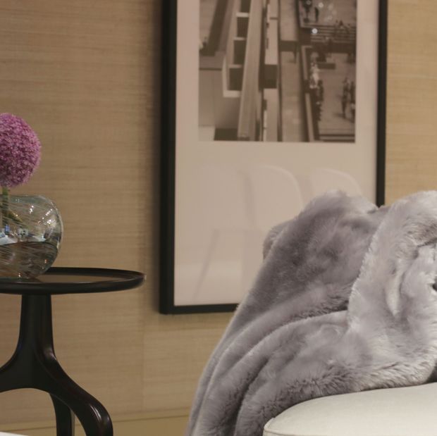 Close-up interior vignette showing a pale upholstered armchair or sofa with a soft grey faux-fur throw, beside a small black pedestal side table holding a glass vase with purple flowers. The warm textured beige wall features a large black-framed monochrome architectural photograph. The styling is refined, cozy, and neutral with soft textural layering.