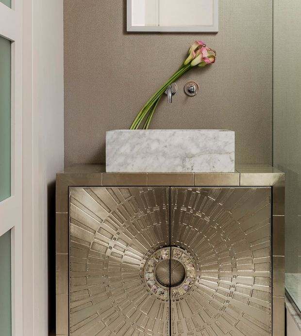 Close-up view of a refined bathroom vanity featuring a sculptural metallic cabinet with a circular sunburst relief pattern, a white marble vessel basin, wall-mounted chrome tapware, and a taupe textured wall. A framed artwork and calla lilies add decorative detail.