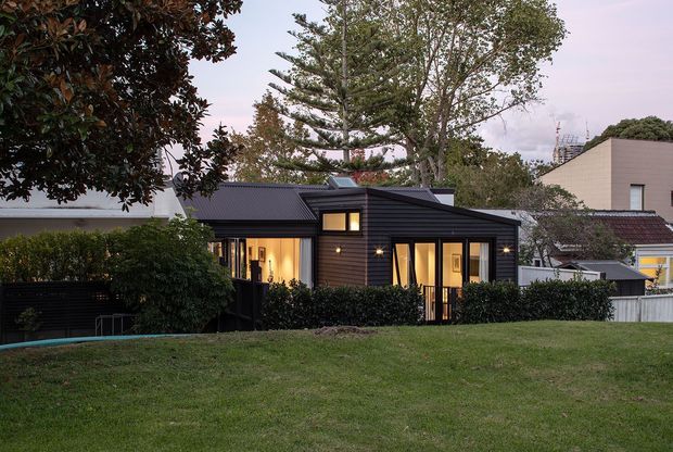 Exterior view of a compact, single-storey dark-clad house set within a suburban garden. The building has a low-pitched, intersecting roof form, horizontal dark wall cladding, extensive warm-lit glazing, and a small raised deck or entry platform. Mature trees, hedges, lawn, and neighbouring homes surround the property.