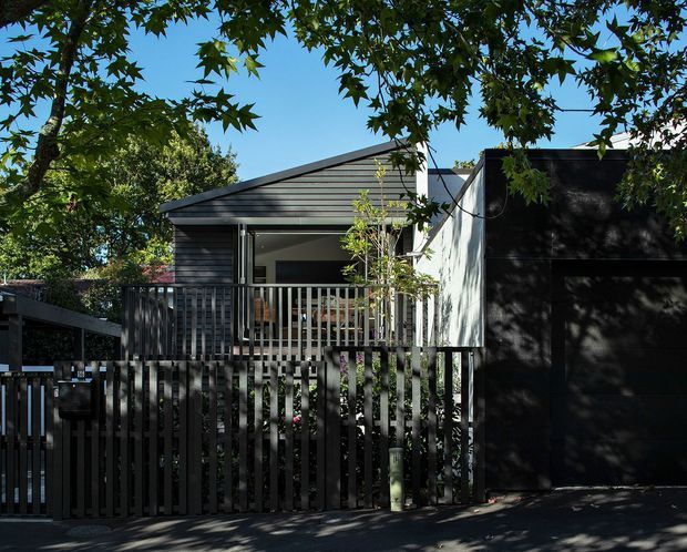 Street-facing view of a compact contemporary house with dark horizontal cladding, a low-pitched roof, and a raised front balcony or deck enclosed by vertical timber battens. A dark slatted fence and gate screen the lower frontage, while mature trees cast dappled shadows across the facade and pavement.
